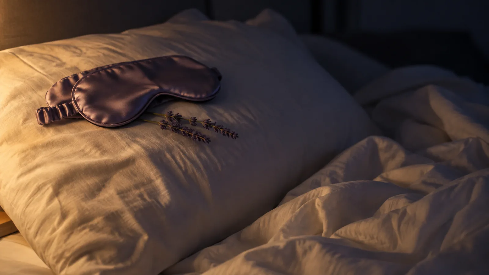 A soft pillow on rumpled white sheets with a lavender eye mask and a sprig of dried lavender in low lamplight.