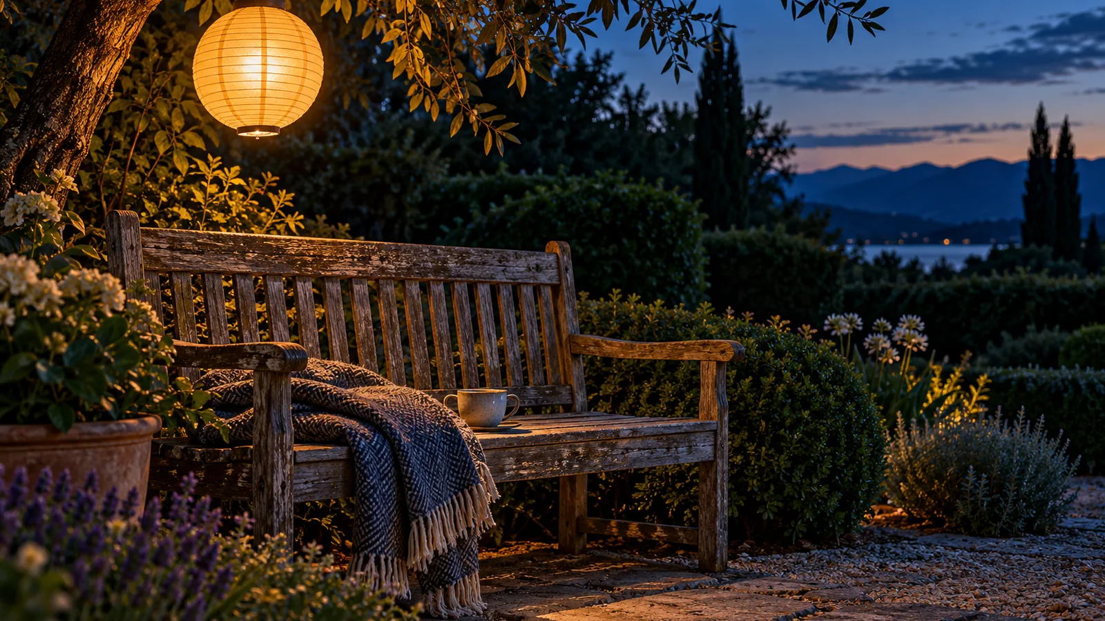 A garden bench at dusk with a folded blanket, a cup and a glowing paper lantern above